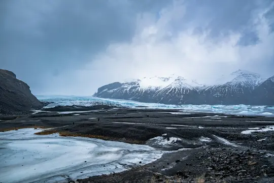 A wide landscape shows a vast glacial plain with dark volcanic rock and patches of ice, leading to a blue glacier and…