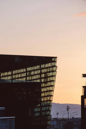 En moderne bygning med en mørk, geometrisk glasfacade er silhuet mod en blød orange og lyserød solnedgangshimmel. Afsides...