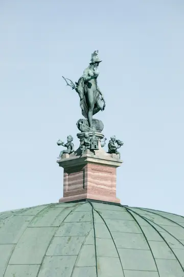 A bronze statue of a helmeted female figure holding a sphere and bow, flanked by three smaller figures, on a striped…