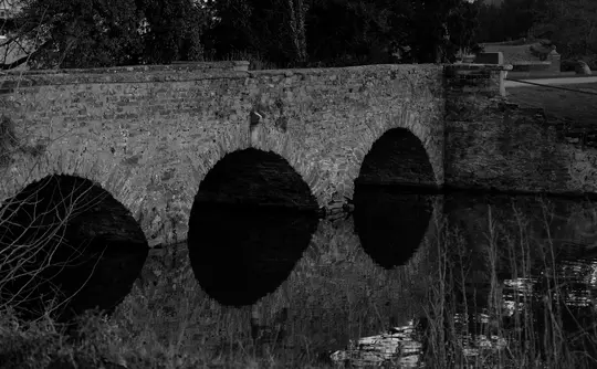 A black and white photo of an old stone bridge with three arches, reflected in the calm water below. Trees are visible…