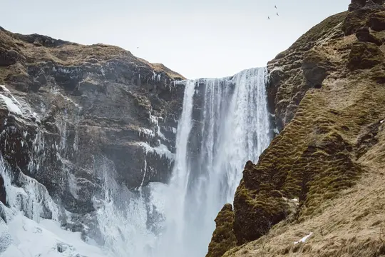 A wide shot of a powerful waterfall plunging down a dark, rocky cliff. The surrounding slopes are covered in brown moss and…