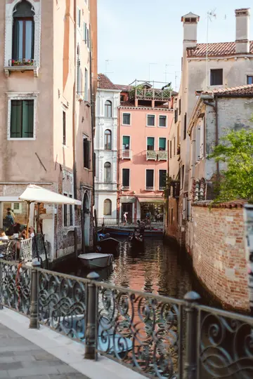En smal kanal i Venedig med historiske bygninger i fersknerøde og cremefarver, flere både og en dekorativ rækværk.