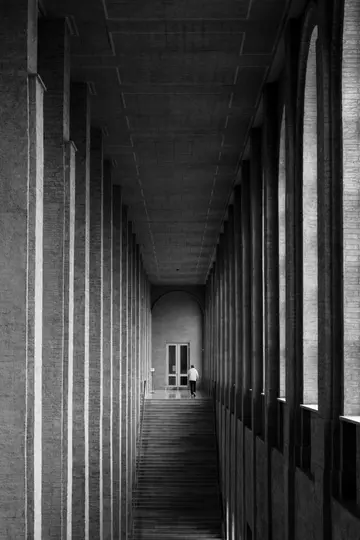 A black and white photo of a person walking up a long staircase in a corridor lined with tall, textured columns.