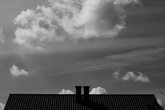A black and white photo of a dark tiled roof with two prominent chimneys silhouetted against a sky filled with clouds.