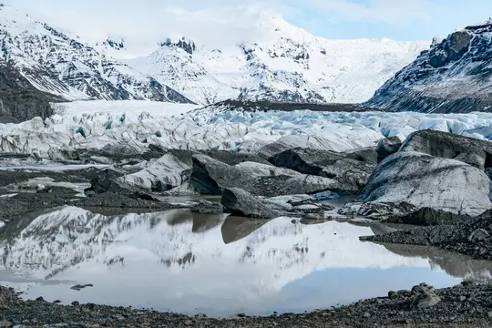 En stor gletsjer med takkede isformationer og snedækkede bjerge under en overskyet himmel, reflekteret i en stille dam.