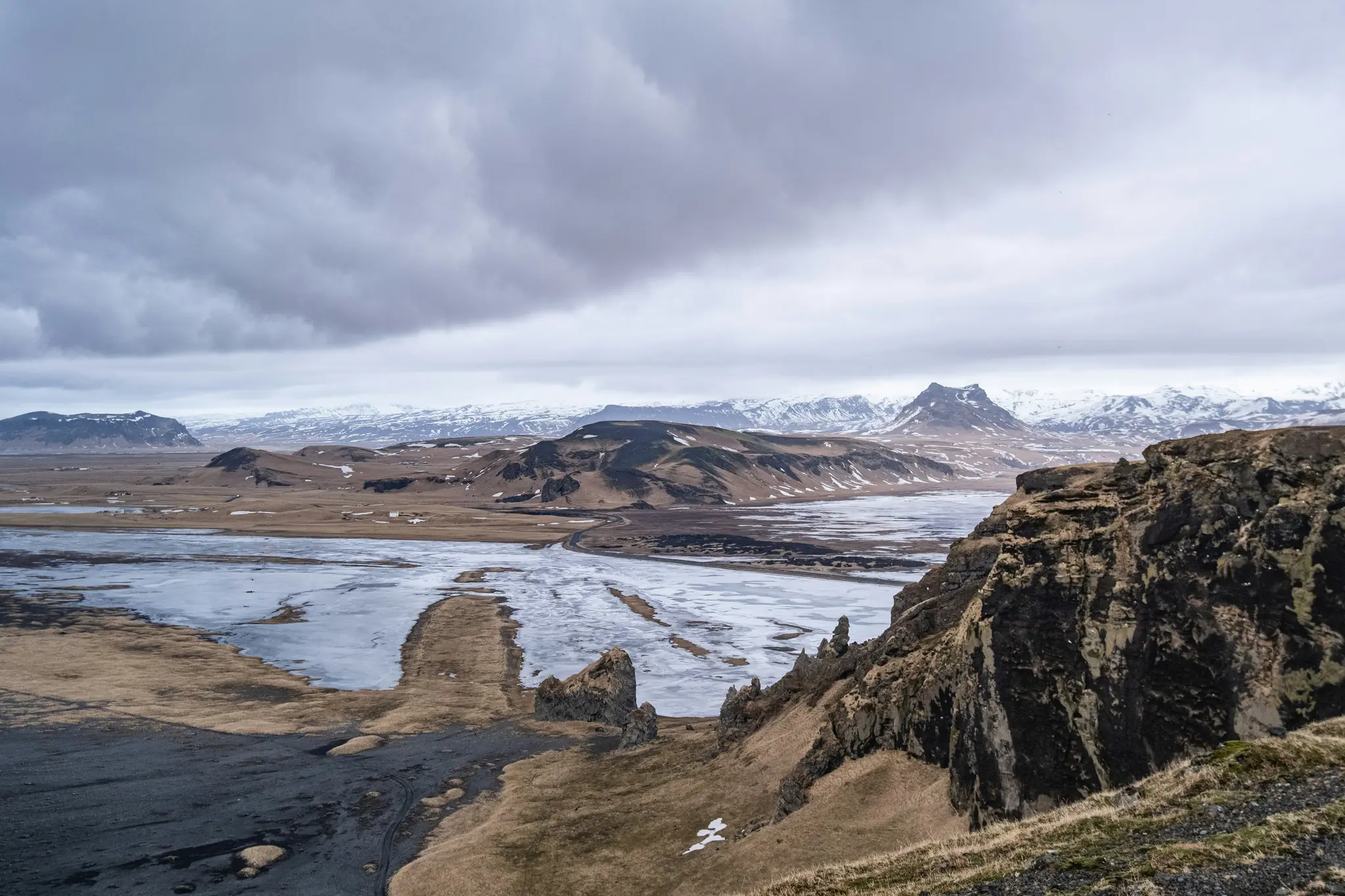 A wide, high-angle landscape photo shows a valley with a partially frozen river, brown hills, and distant snow-capped…
