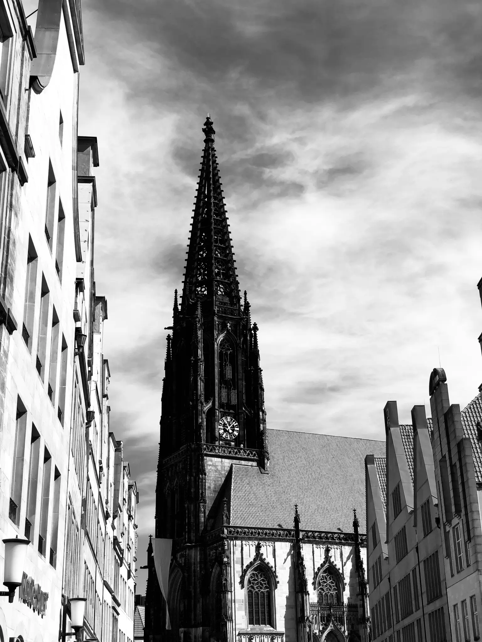 A dramatic black and white, low-angle shot of a towering gothic church spire with a clock, flanked by historic buildings.