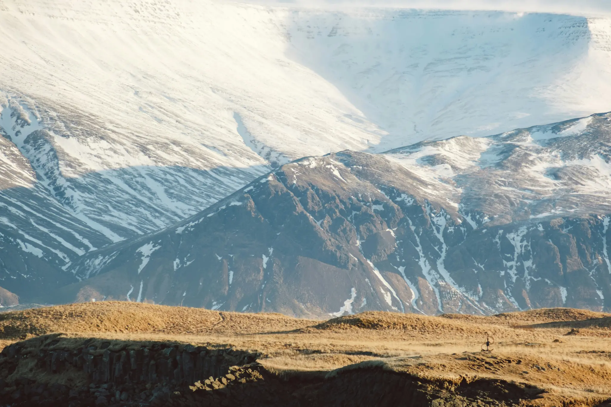 A wide landscape photo showing a range of snow-covered mountains under a bright sky, with golden-brown grassy hills in the…
