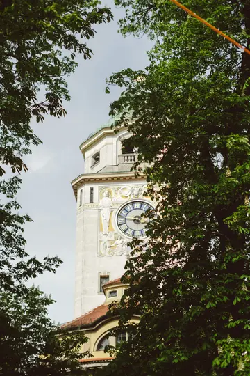 A detailed view of an ornate clock tower with a green dome, partially obscured by vibrant green tree leaves.