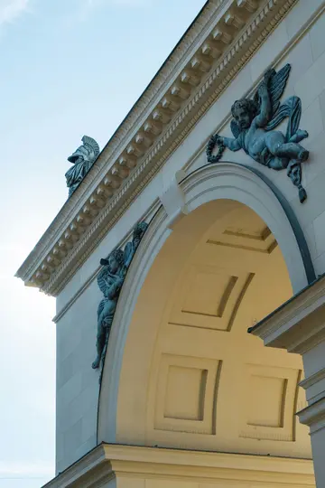 A low-angle shot of a cream-colored building with ornate architectural details, including two cherub sculptures and a…