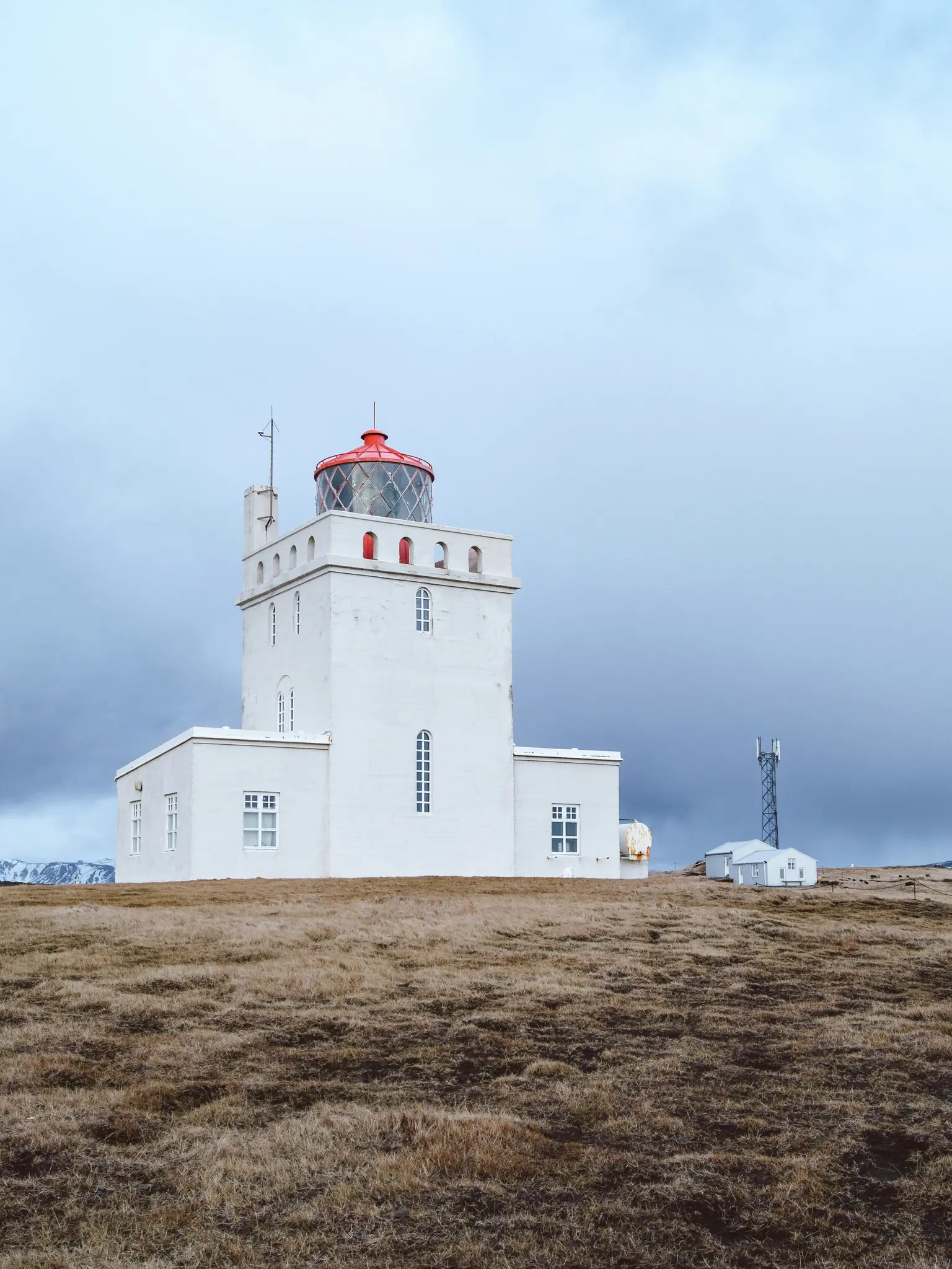 A tall, white lighthouse with a bright red lantern room stands prominently on a brown, grassy hill under a cloudy, grey…
