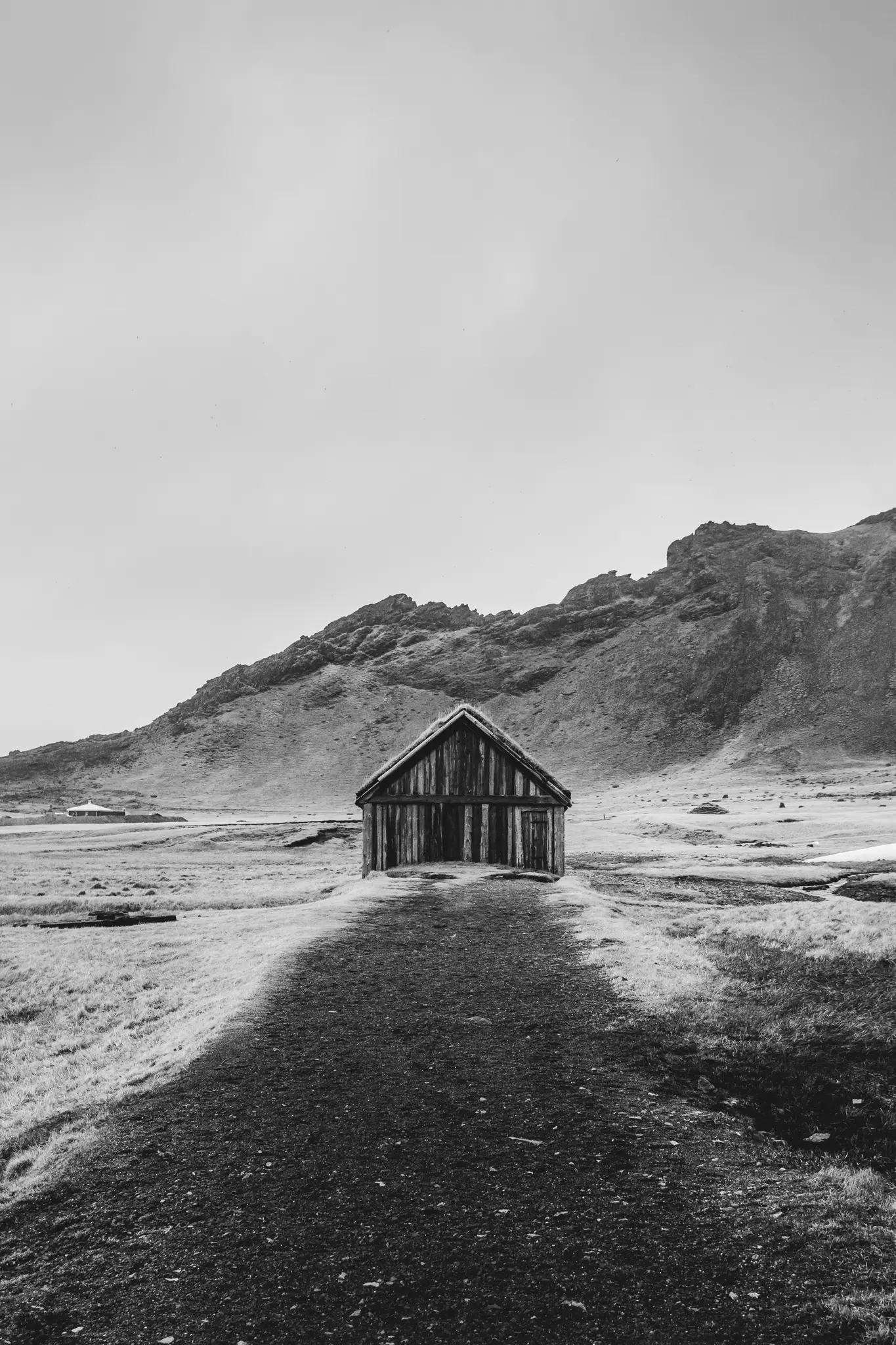 A black and white photo shows a small, weathered wooden hut with a grass roof at the end of a dirt path in a vast, empty…