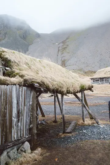 A weathered wooden building with a thick, dry grass roof stands in the foreground. Behind it, a rocky, grey mountain is…