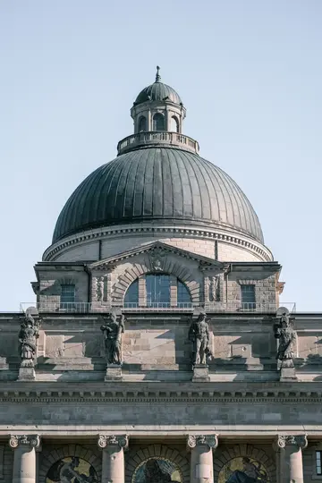 En detaljeret visning af den øverste del af en klassisk stenbygning med en stor, mørk kuppel og en udsmykket facade med