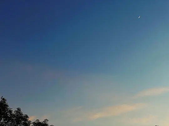 A thin crescent moon hangs in a vast blue sky, transitioning to lighter blue and faint orange clouds near the horizon. Dark…