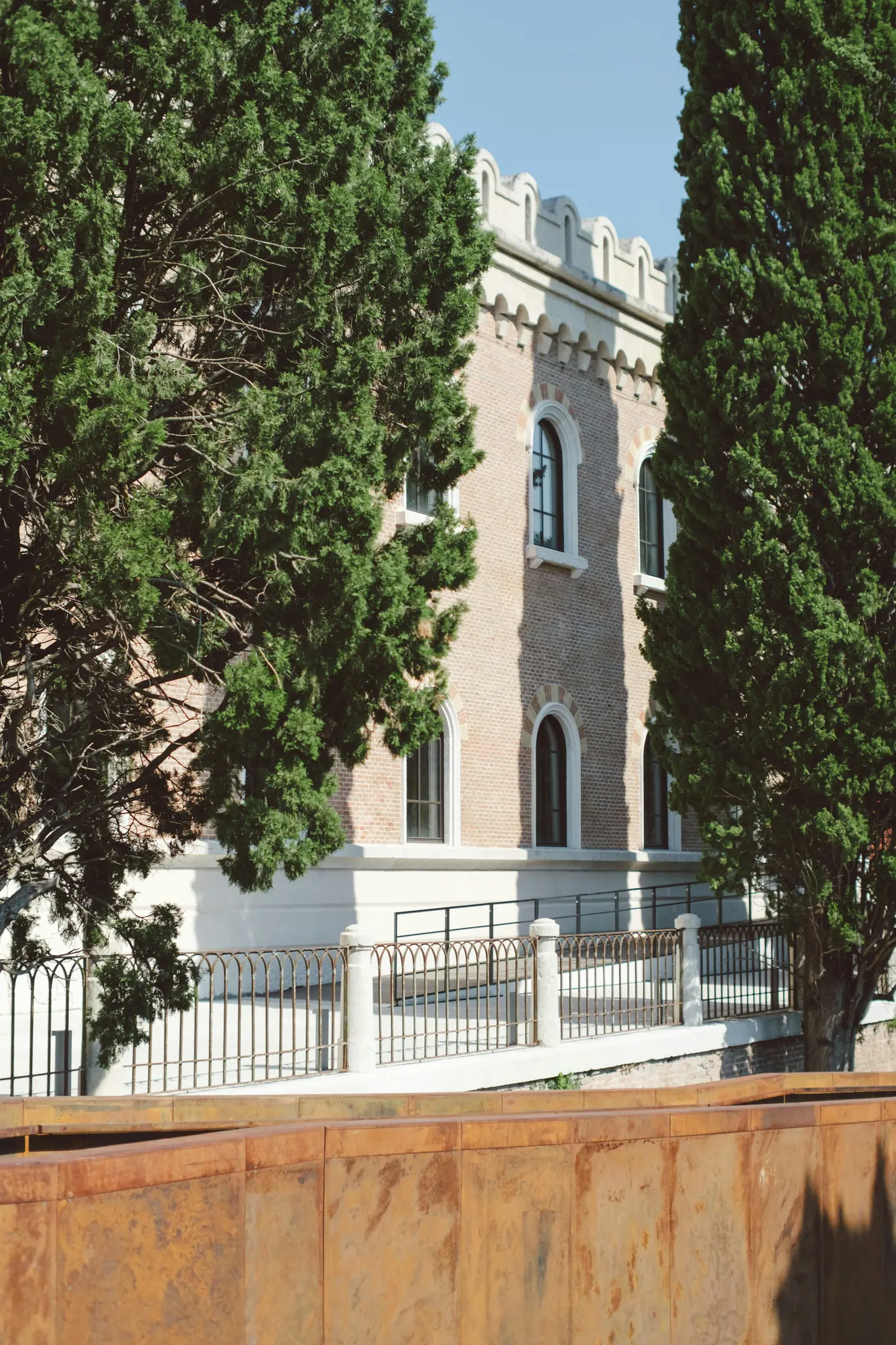 A light brown brick building with arched windows and a decorative white roofline is framed by tall green trees. A rusty…