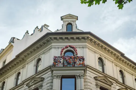 A low-angle shot of a cream-colored building's upper floors, featuring an ornate corner balcony adorned with pink, purple…