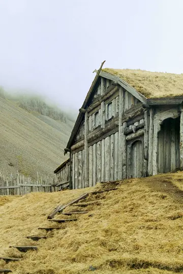 Et vejrbidt træhus med et grønt græstag står på en gylden græsklædt bakke. Et stejlt, stenet bjerg er bagved.