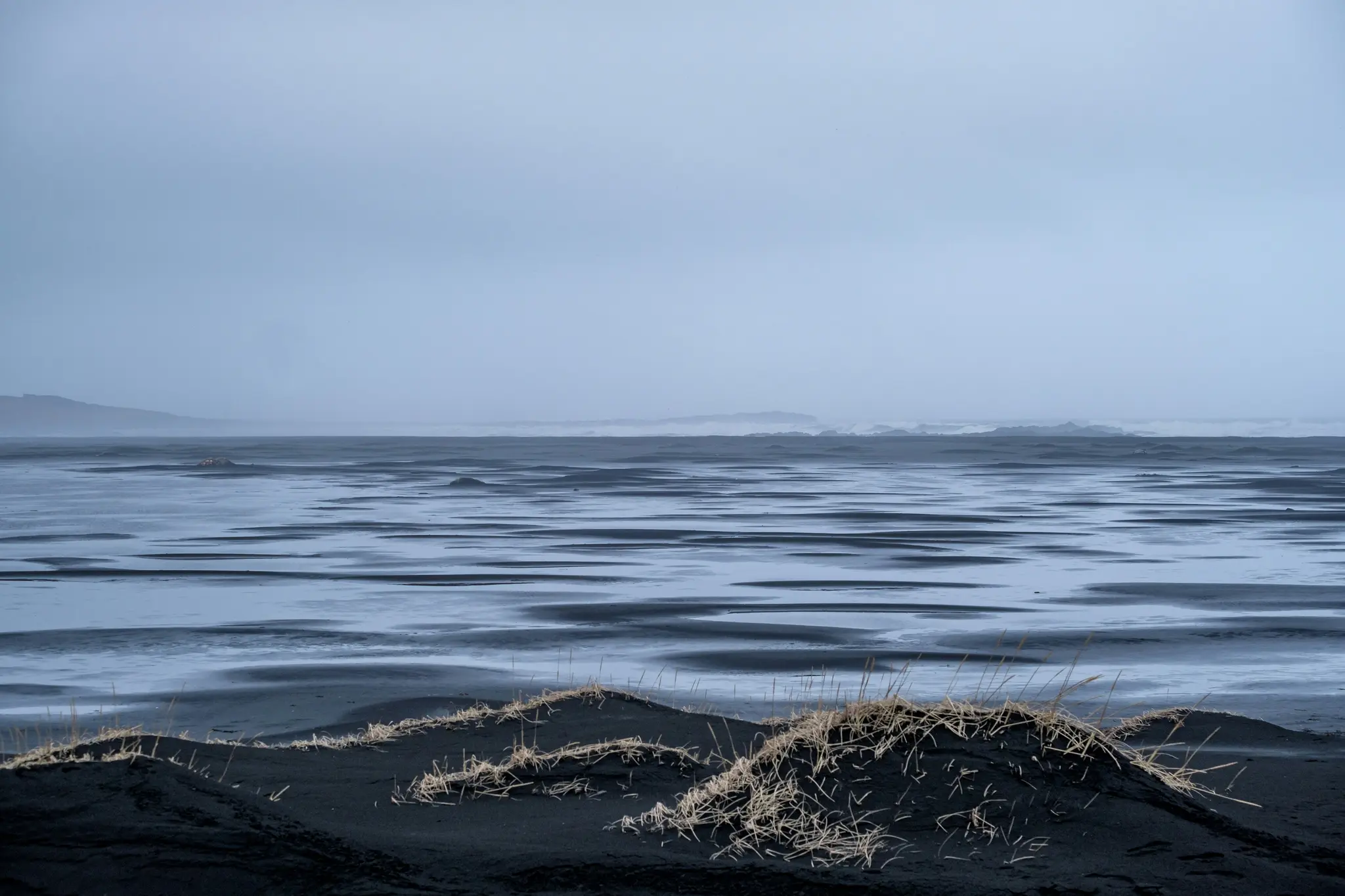 A wide landscape photo of dark, possibly black, sand flats with numerous shallow, reflective water pools under a grey…
