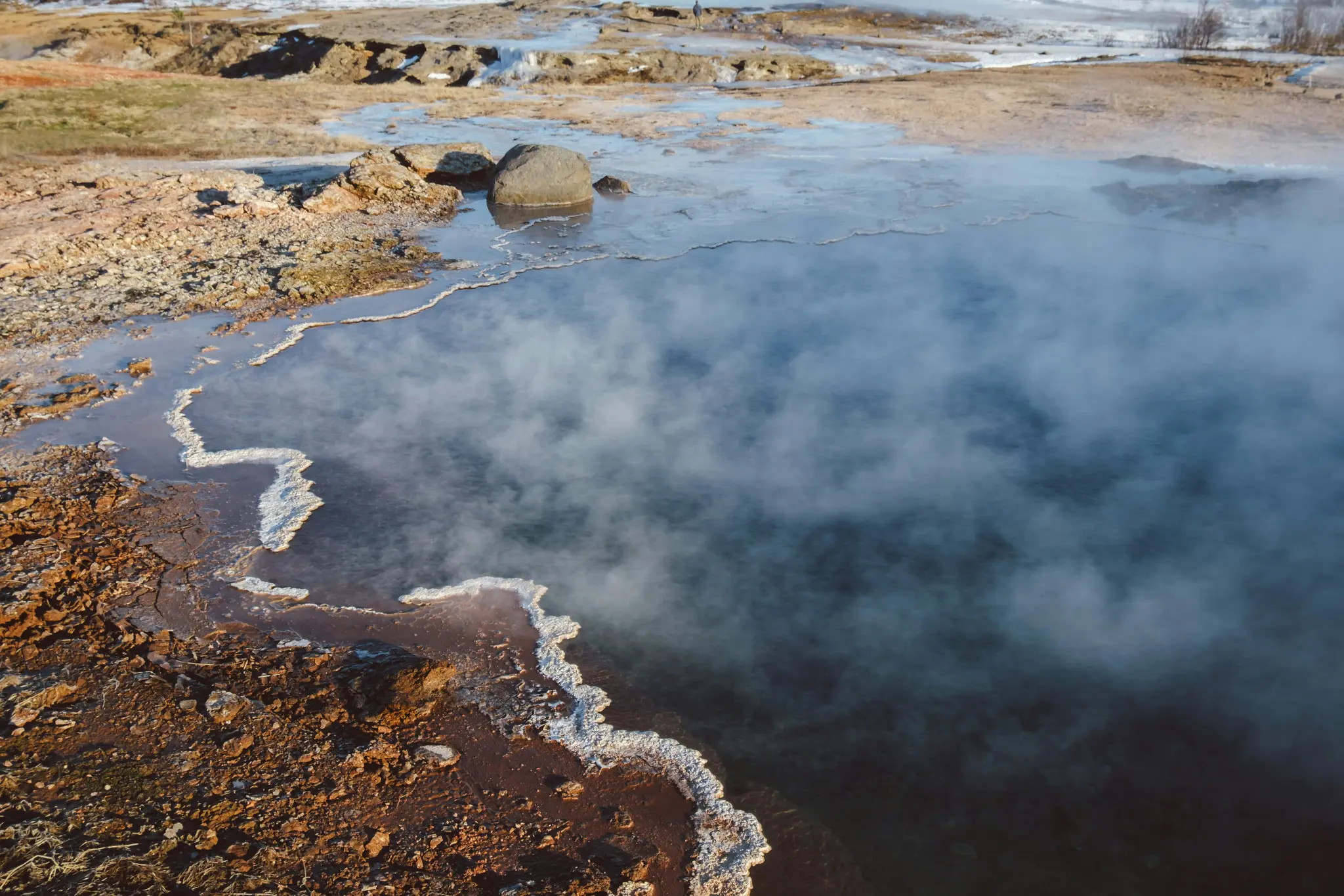 A high-angle view of a steaming, dark blue-green geothermal pool. The water's edge is a white, wavy mineral deposit against…