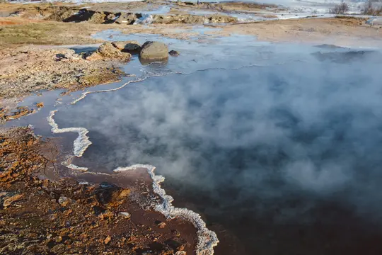 A high-angle view of a steaming, dark blue-green geothermal pool. The water's edge is a white, wavy mineral deposit against…