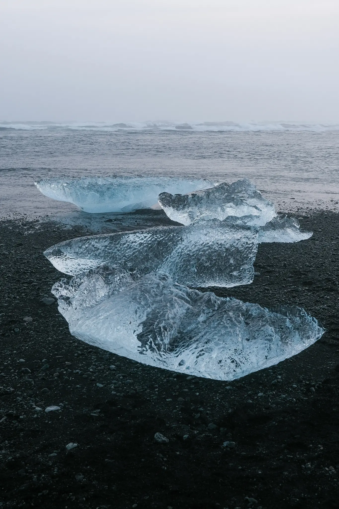 Several clear blue ice chunks rest on a black pebble beach, with gray ocean waves and an overcast sky in the background.
