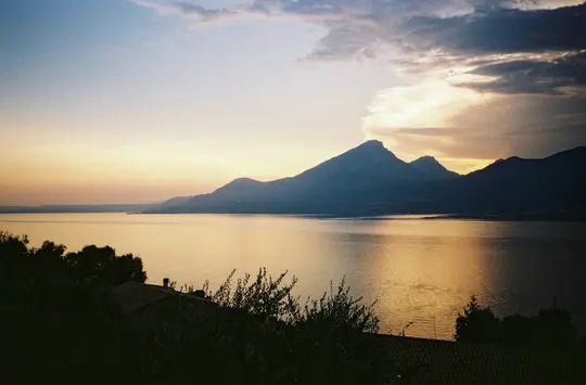 A wide landscape photo of a lake reflecting the golden light of a sunset, with dark mountains silhouetted in the distance.…