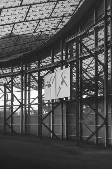 A black and white photo of a building's interior, featuring a glass roof and metal beams. Two white square signs with black…