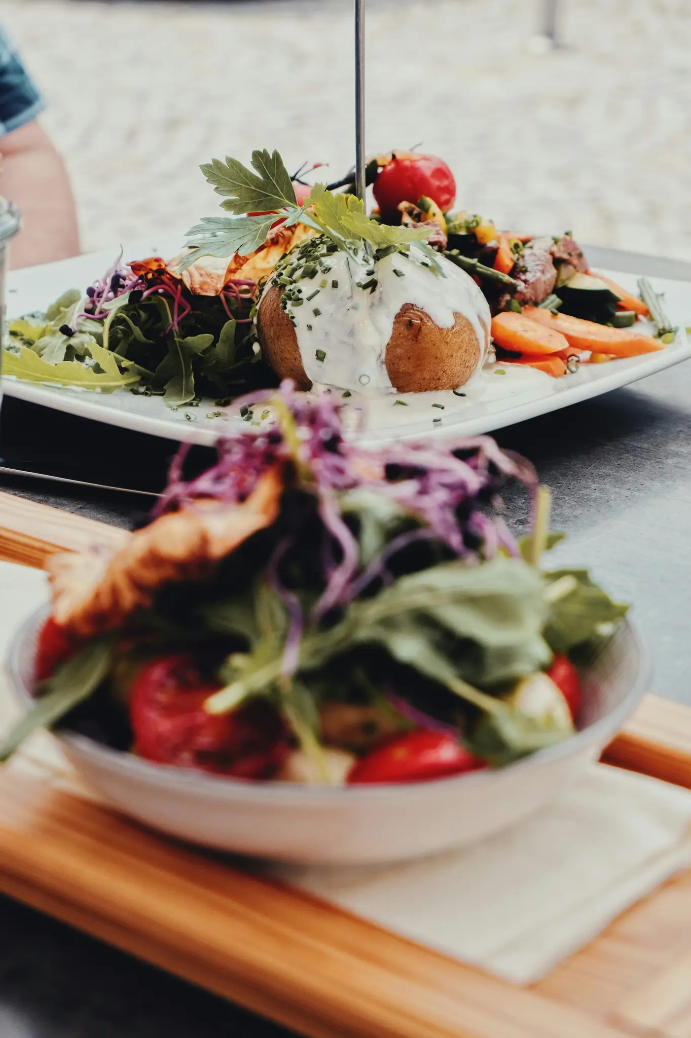A baked potato topped with white sauce and chives, surrounded by a colorful mix of vegetables and a side salad, on a white…