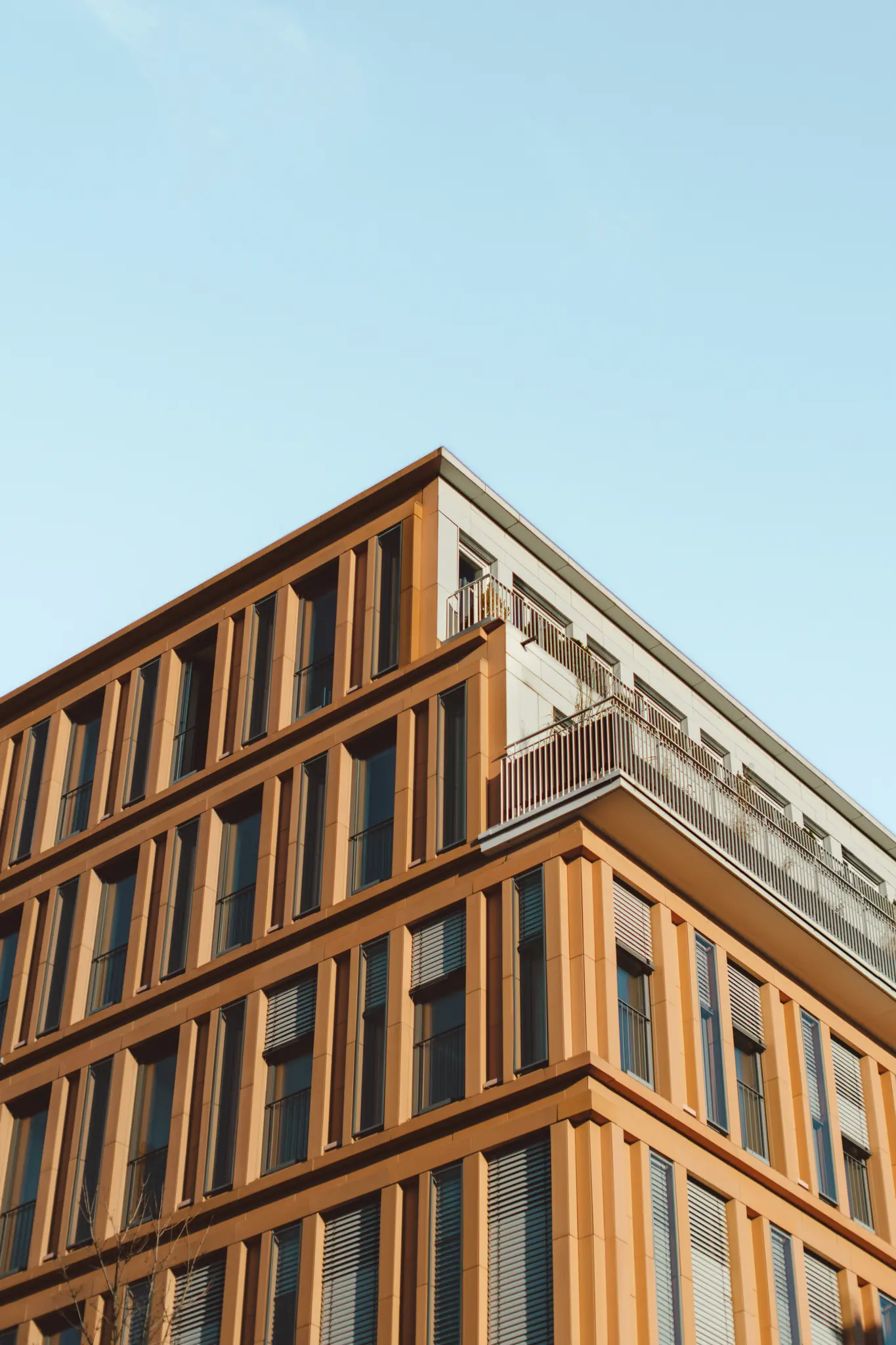 A low-angle view of a modern building with an orange-brown facade, numerous windows, and balconies, set against a clear…