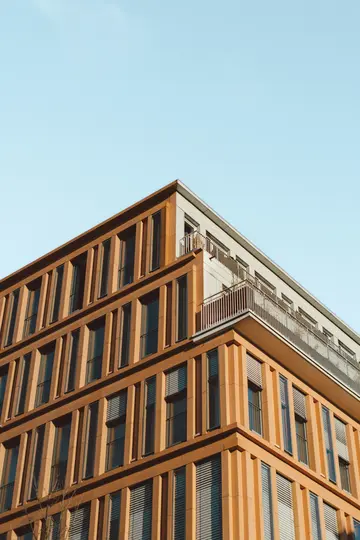 A low-angle view of a modern building with an orange-brown facade, numerous windows, and balconies, set against a clear…