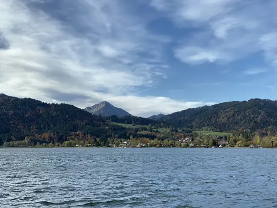 A wide landscape photo of a lake with ripples on its surface, reflecting a partly cloudy sky. On the far shore, a village…