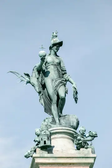 A weathered bronze statue of a woman in a helmet, holding an orb and a wreath, with a deer head and cherubs.