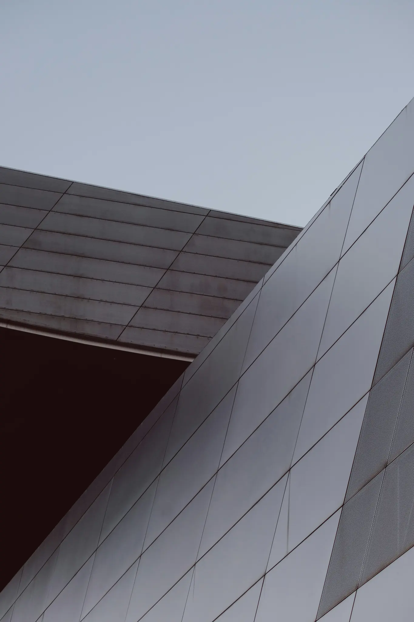 An abstract, low-angle view of a modern building with grey and dark brown geometric panels against a light blue sky.