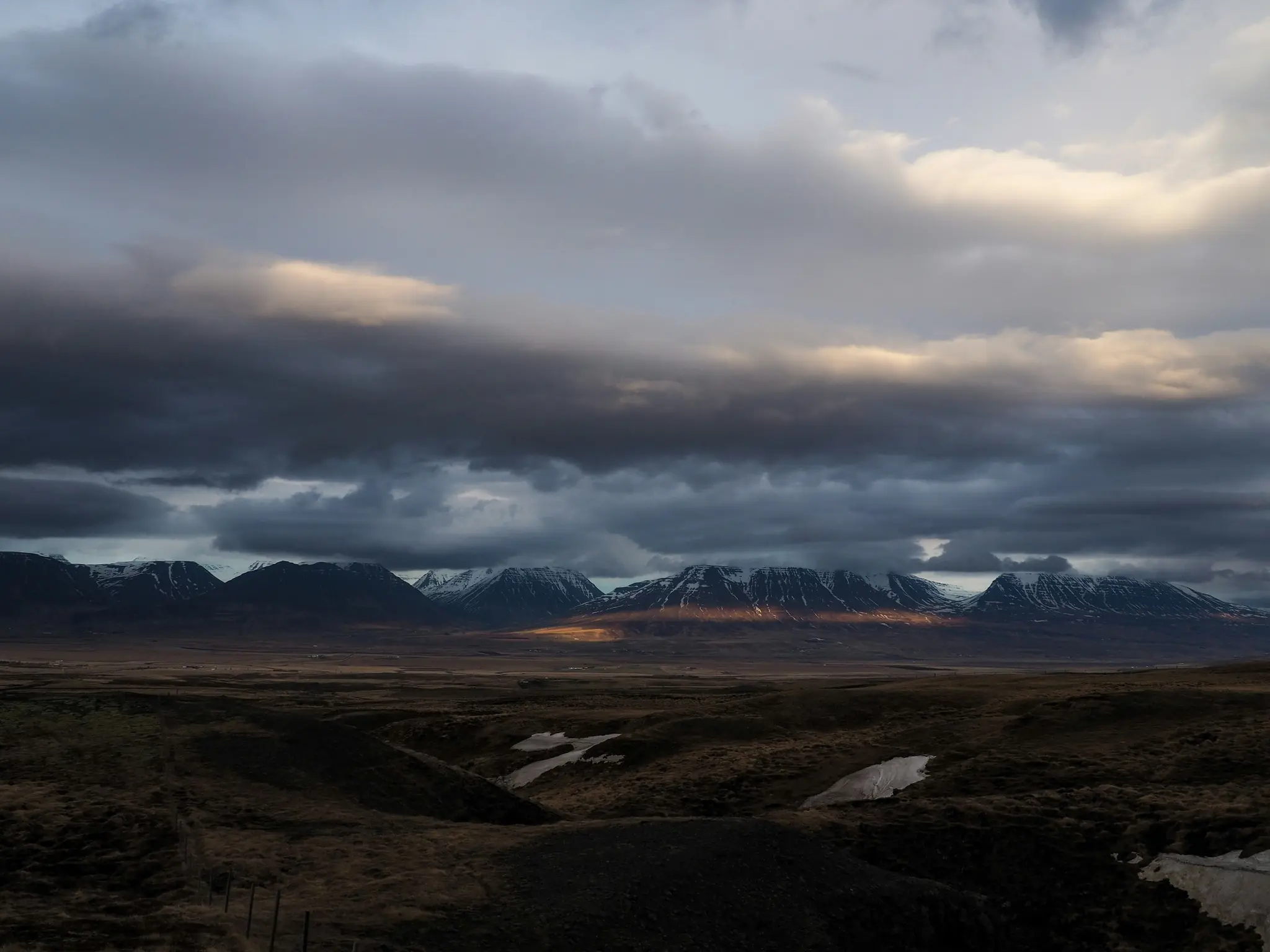 Ett brett landskapsfoto visar en rad mörka, snötäckta berg under ett tungt, mörkt molntäcke. Ett smalt band…
