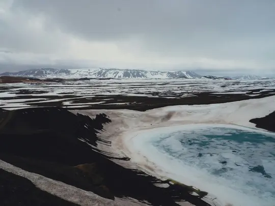 En flygvy över en vulkanisk krater i med isblått vatten, omgiven av mörk jord och snöfläckar. Två små…