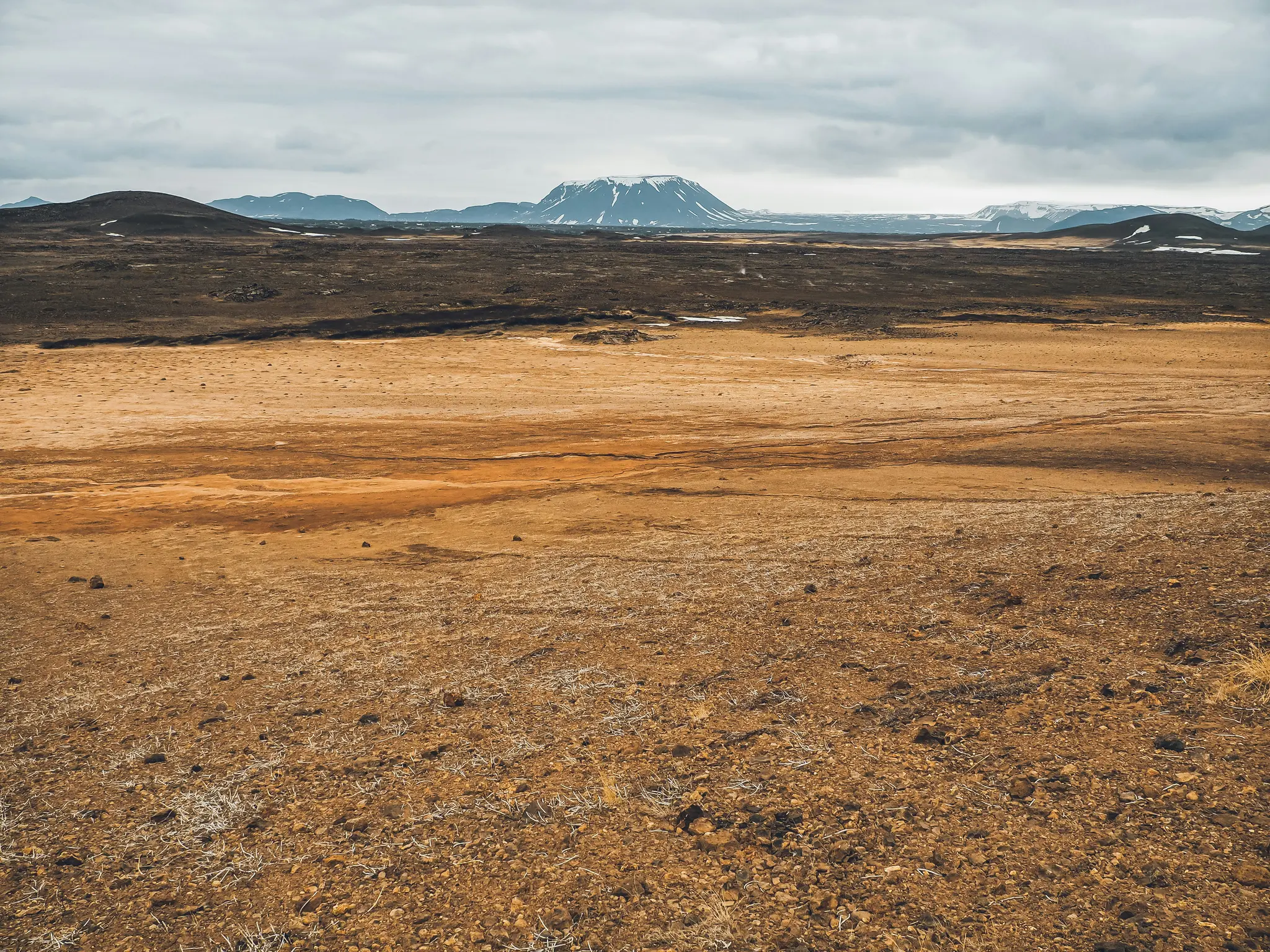 En vidsträckt vy över ett öde, stenigt landskap. Förgrunden består av rödbrun jord med sparsam torr vegetation. I...