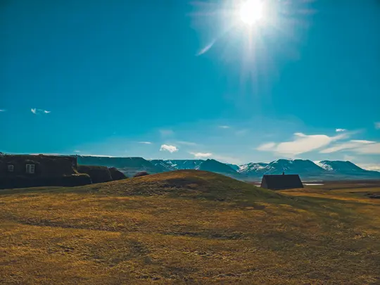 Ett brett landskapsfoto som visar två byggnader med torvtak på gräsbevuxna kullar i förgrunden, med snötäckta berg i