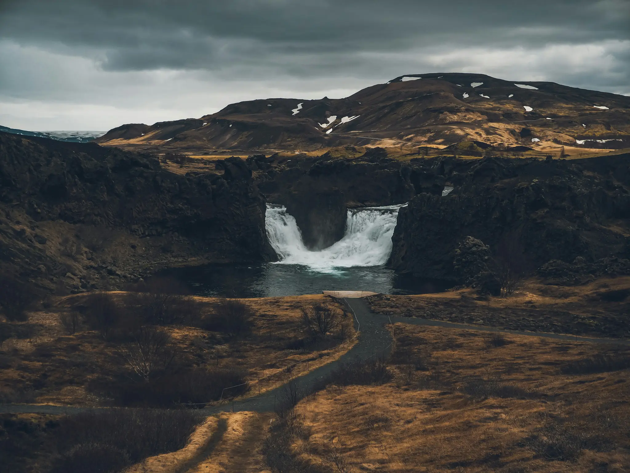 Ett brett landskapsfoto visar ett kraftfullt vattenfall som störtar ner i en mörk damm, omgivet av mörkbruna och gröna