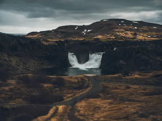 Ett brett landskapsfoto visar ett kraftfullt vattenfall som störtar ner i en mörk damm, omgivet av mörkbruna och gröna