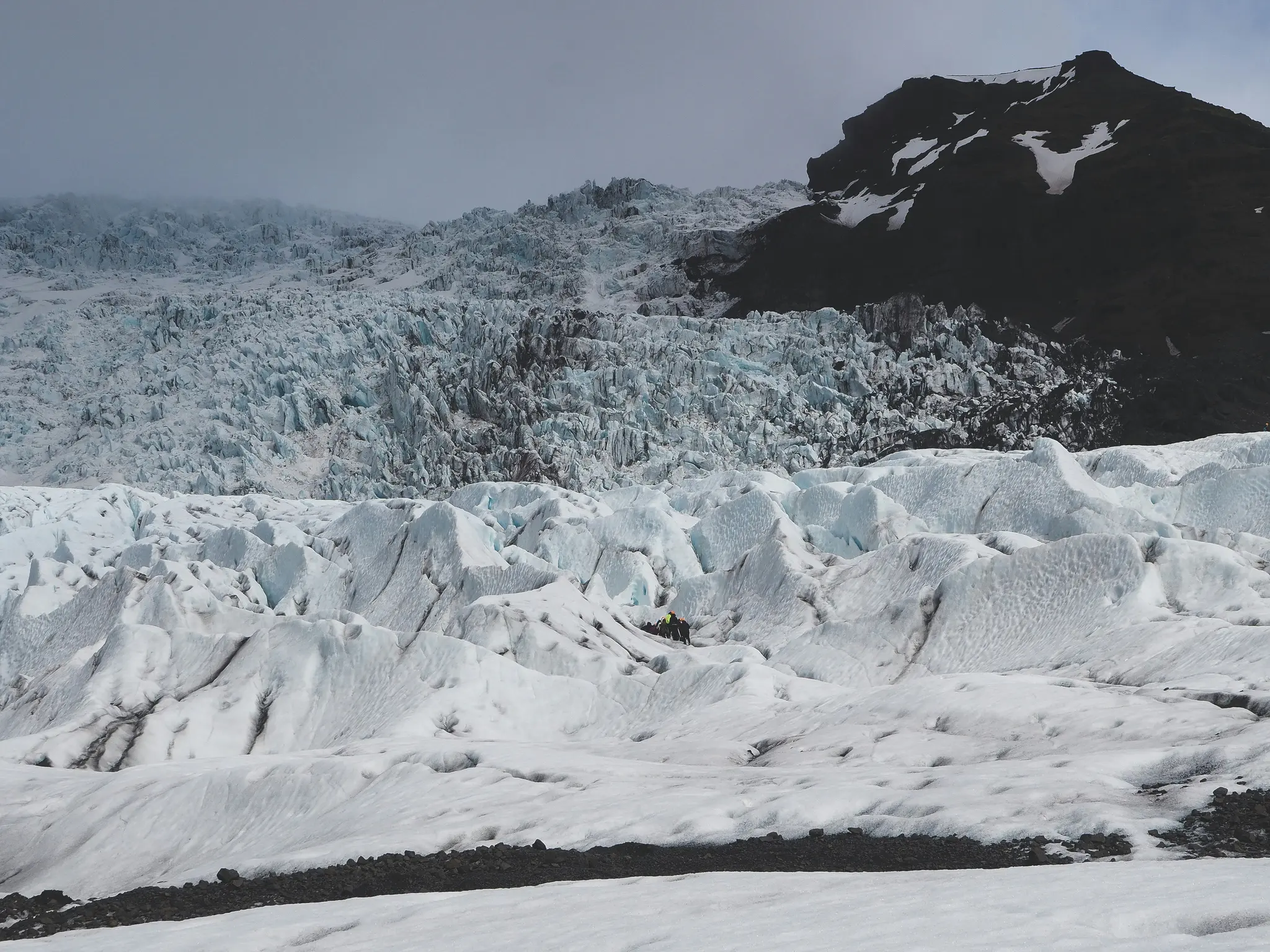 En vid bild av en glaciär med taggiga isformationer i vitt och ljusblått, och ett mörkt, klippigt berg bakom.