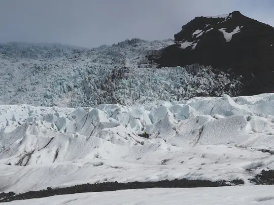 En vid bild av en glaciär med taggiga isformationer i vitt och ljusblått, och ett mörkt, klippigt berg bakom.