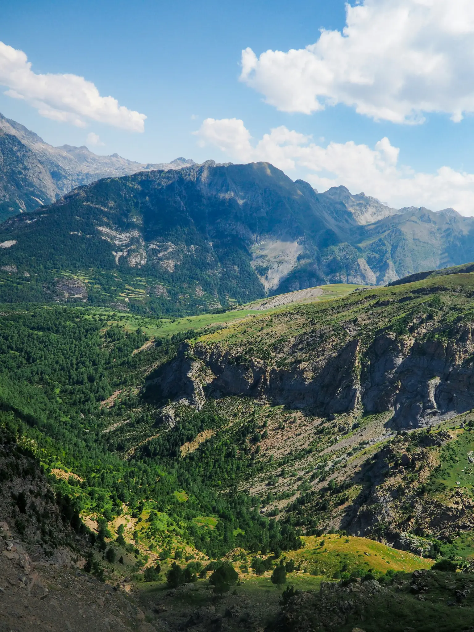 Ett brett landskapsfoto som visar en vidsträckt bergregion med djupa dalar täckta av täta gröna skogar och klippiga