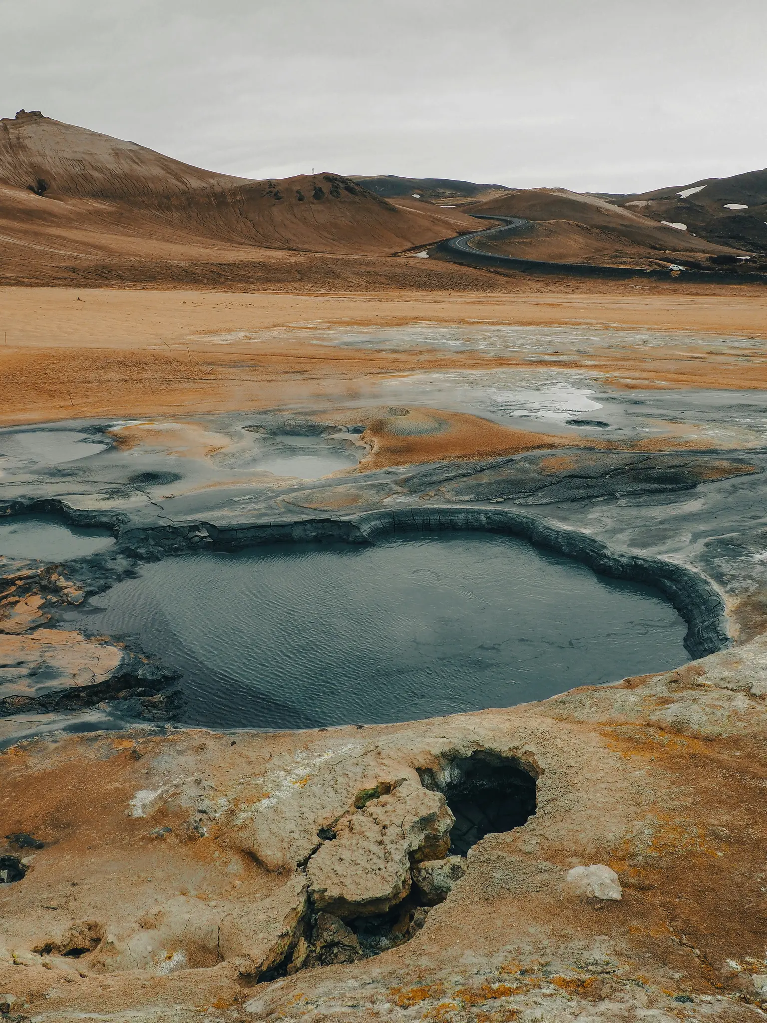 En bred bild av ett geotermiskt landskap med en mörk, stilla vattenpool i förgrunden, omgiven av sprucken...