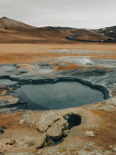 En bred bild av ett geotermiskt landskap med en mörk, stilla vattenpool i förgrunden, omgiven av sprucken...