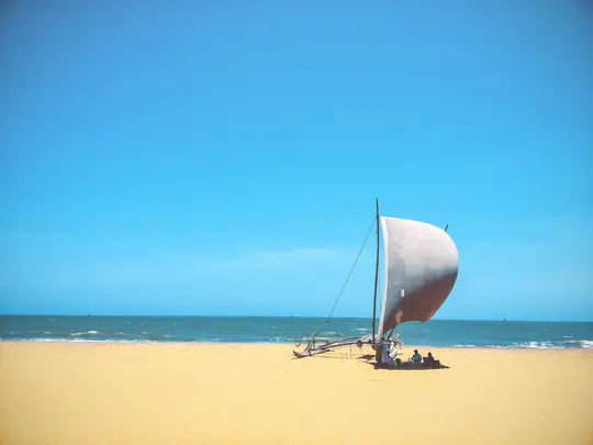 En traditionell segelbåt med ett stort vitt segel vilar på en bred, gyllene sandstrand. Havet är lugnt med små vågor.