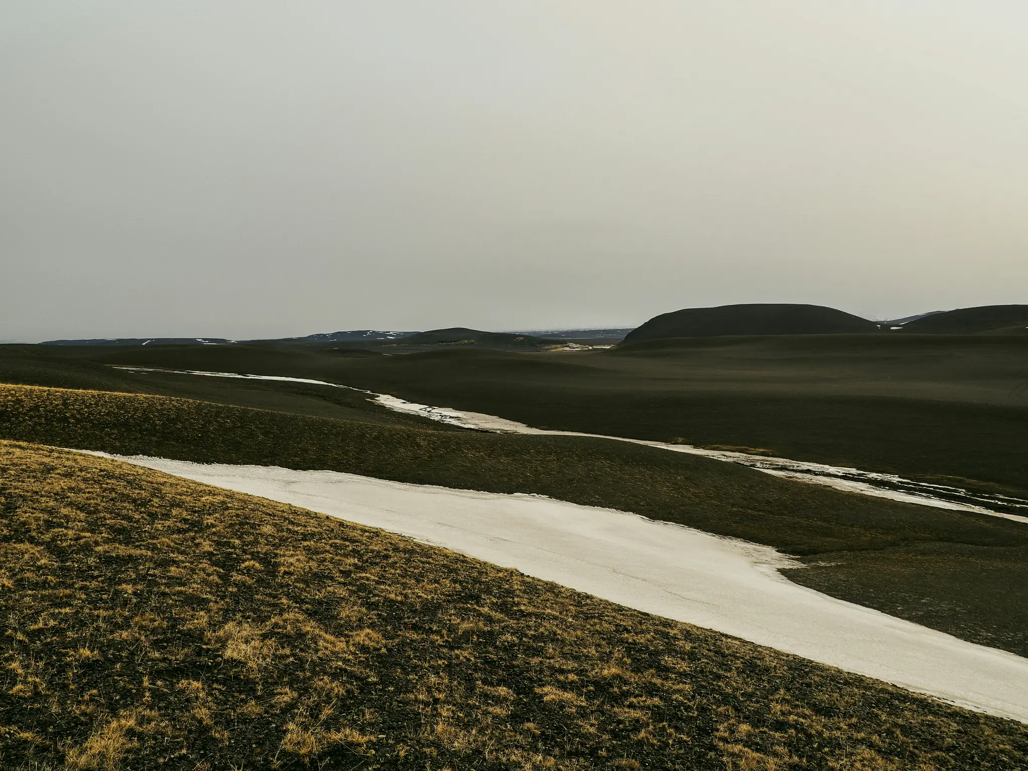 Ett brett landskapsfoto som visar böljande kullar täckta av mörkt, torrt gräs och fläckar av vit snö under en grå himmel.