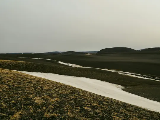 Ett brett landskapsfoto som visar böljande kullar täckta av mörkt, torrt gräs och fläckar av vit snö under en grå himmel.