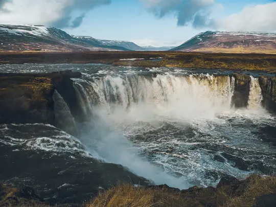 Ett brett landskapsfoto av ett stort, kraftfullt vattenfall med vitt, skummande vatten som kraschar ner i en mörk, turbulent