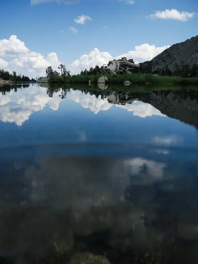 Ett fotografi i delad vy av en lugn bergssjö. Ovanför vattnet reflekteras vita moln på den blå ytan. Berg omger sjön.